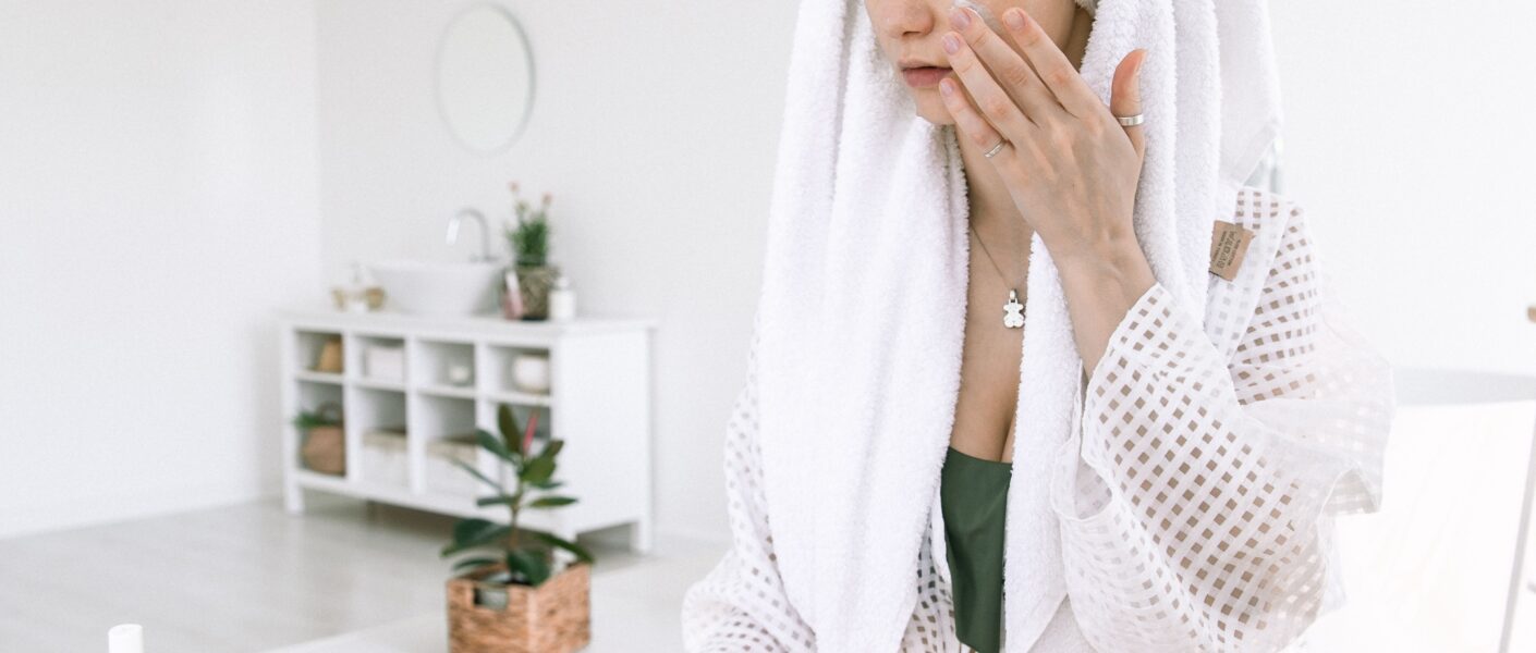 Woman in bathrobe applying skincare in a modern bathroom setting with beauty products.